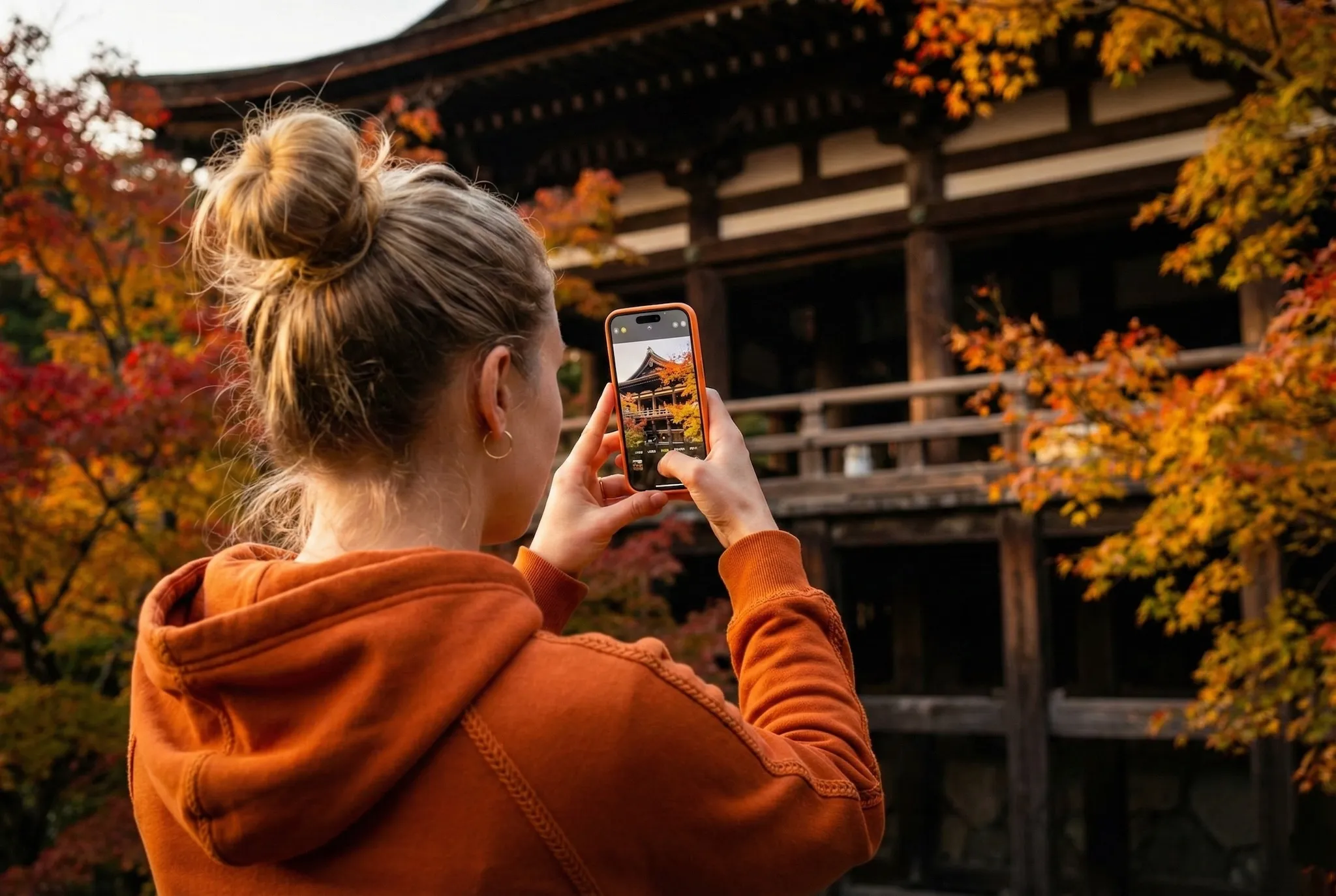 Rear view of a woman in an orange hoodie taking a photo with her smartphone of a wooden Japanese temple amidst vibrant autumn foliage.